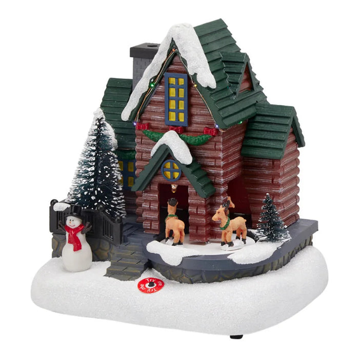 Red log house scene with a green roof, reindeer, a snowman and snowy trees on a white base