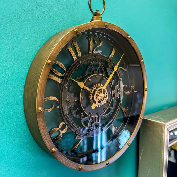 Industrial-style wall clock with a vintage brass-look finish and faux internal gears, hanging on a teal wall beside a mirror, styled in a showroom setting.