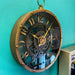Industrial-style wall clock with a vintage brass-look finish and faux internal gears, hanging on a teal wall beside a mirror, styled in a showroom setting.