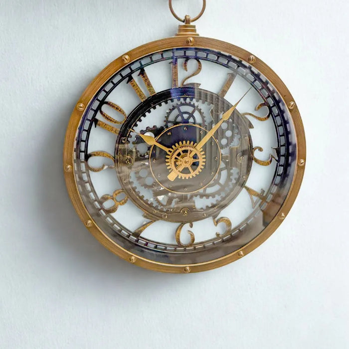 Industrial-style wall clock with a vintage brass-look finish and faux internal gears, hanging on a teal wall beside a mirror, styled in a showroom setting.