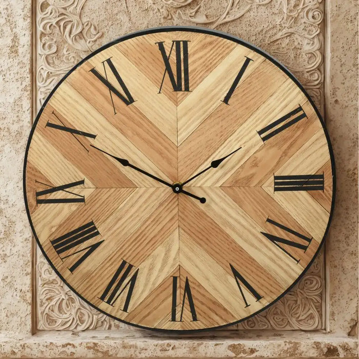 Chevron-patterned MDF clock with Roman numerals, styled on a round gold table against a dark feature wall with abstract artwork and modern glass wall lights.