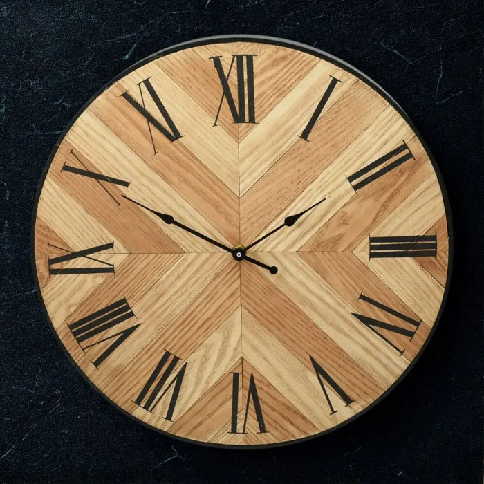 Chevron-patterned MDF clock with Roman numerals, styled on a round gold table against a dark feature wall with abstract artwork and modern glass wall lights.