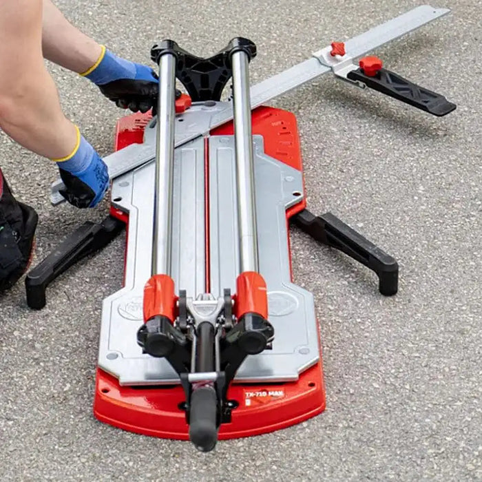 Rubi TX-710 MAX manual tile cutter with red handle, steel guide rails, and side arms, placed on a workshop surface.