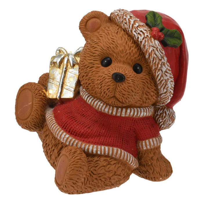 Sitting Christmas bear in a Santa hat and red jumper holding a gold wrapped gift