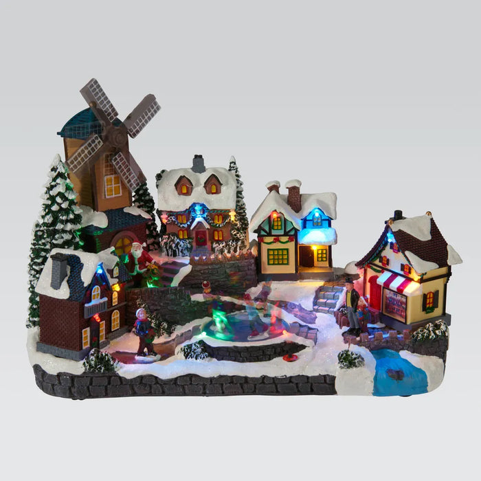Lit snowy village scene with a windmill, cottages and a sweet shop around an ice rink with skaters